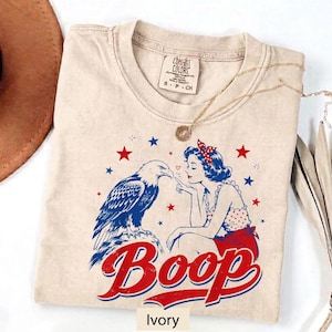 May include: An ivory t-shirt featuring a graphic print of a bald eagle and a person with a red polka dot hair accessory. The design includes red and blue stars and the text Boop in red and blue lettering. The shirt has a crew neckline.