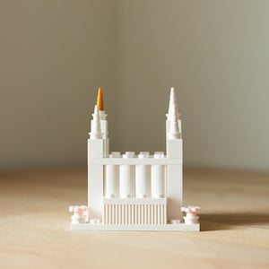May include: A white Lego model of a building with two tall spires and a gold-colored top. The building has a series of vertical columns and a detailed facade. The model is set on a wooden surface.