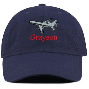 May include: A navy blue baseball cap featuring an embroidered gray airplane graphic positioned above the red embroidered text Grayson. The cap has a structured crown, a curved brim, and a button at the top center of the fabric dome.