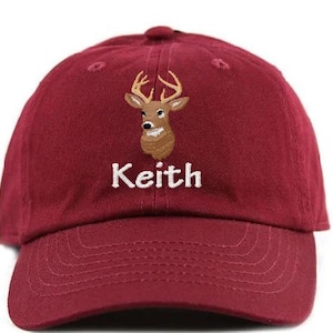 May include: A maroon baseball cap with a curved brim. The cap features an embroidered deer head with antlers and the name "Keith" in white block letters. The cap is made of a soft fabric and has a top button.