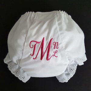 May include: White cotton baby bloomers with a pink embroidered letter "M" and the initials "NL". The bloomers have a white lace trim.