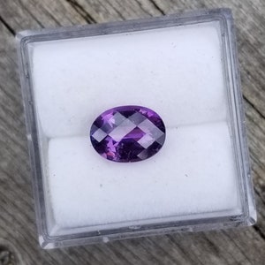 May include: A purple oval gemstone with facets in a clear plastic box.