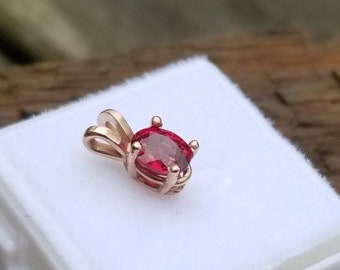 May include: A red gemstone pendant in a gold setting, displayed in a white box.
