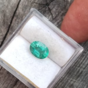 May include: A vibrant green oval emerald gemstone in a small white box.