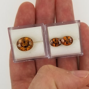 May include: Two oval-shaped gemstones in clear plastic boxes. The gemstones are a deep orange color with a slight reddish hue.