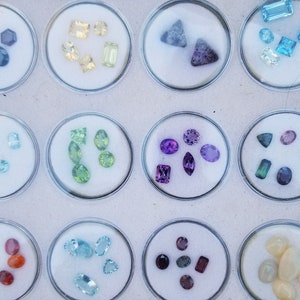 May include: A collection of small, colorful gemstones displayed in clear plastic containers. The gemstones are arranged in rows and include various shapes, sizes, and colors, such as purple, green, blue, and red.