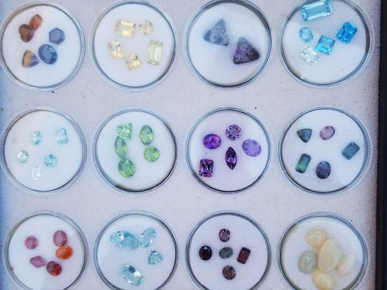 May include: A collection of small, colorful gemstones displayed in clear plastic containers. The gemstones are arranged in rows and include various shapes, sizes, and colors, such as purple, green, blue, and red.