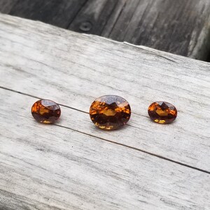 May include: Three oval-shaped gemstones, ranging in size, with a warm, amber-like color. The stones are set against a light wood background.
