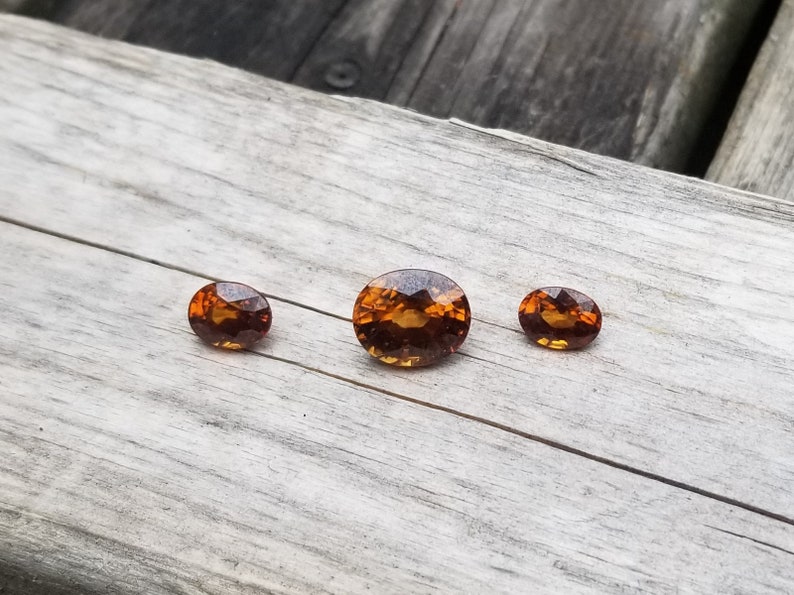 May include: Three oval-shaped gemstones, ranging in size, with a warm, amber-like color. The stones are set against a light wood background.