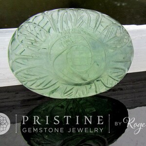 Pode incluir: Um caboch&atilde;o de pedra preciosa verde com um design esculpido. A pedra preciosa tem forma oval e uma cor verde clara. A escultura &eacute; intrincada e apresenta um padr&atilde;o floral.