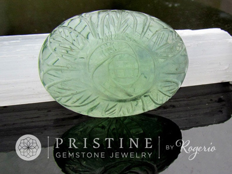Pode incluir: Um caboch&atilde;o de pedra preciosa verde com um design esculpido. A pedra preciosa tem forma oval e uma cor verde clara. A escultura &eacute; intrincada e apresenta um padr&atilde;o floral.