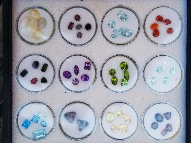 May include: A collection of gemstones displayed in small round containers. The gemstones are arranged by color, with each container holding a different variety of stones. The colors include red, orange, yellow, green, blue, purple, and white.