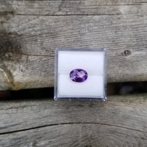 May include: A purple oval gemstone in a clear plastic box