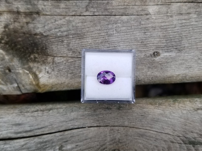 May include: A purple oval gemstone in a clear plastic box