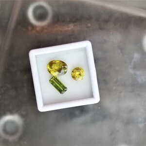 May include: Three green gemstones in a white box. The gemstones are different shapes and sizes. The largest gemstone is oval shaped, the second largest is round, and the smallest is a long rectangle.