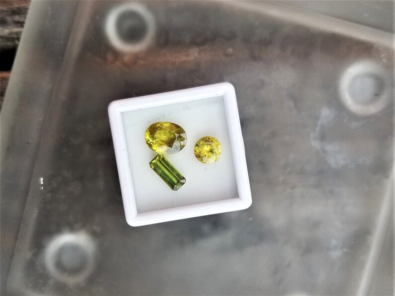 May include: Three green gemstones in a white box. The gemstones are different shapes and sizes. The largest gemstone is oval shaped, the second largest is round, and the smallest is a long rectangle.