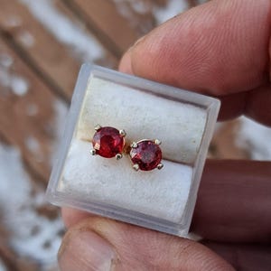 May include: A pair of stud earrings featuring round, deep red gemstones set in gold. The earrings are presented in a clear plastic case with a white interior, against a blurred wooden backdrop.