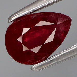 Könnte beinhalten: Ein birnenförmiger, facettierter Rubin, gehalten von einer silbernen Pinzette. Der Rubin hat eine tiefe rote Farbe mit weißen, dreieckigen Facetten. Ein poliertes, geschliffenes Juwel.