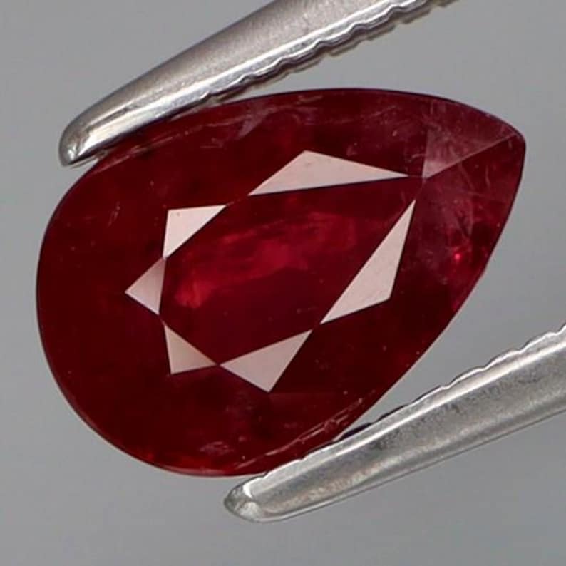 May include: A pear-shaped, faceted ruby gemstone held by silver tweezers. The ruby is a deep red color with white, triangular facets. The gemstone is a polished, cut jewel.