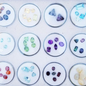 May include: A collection of 12 small glass containers, each filled with a variety of colorful gemstones. The gemstones are arranged in a grid pattern, with each container containing a different type of gemstone.