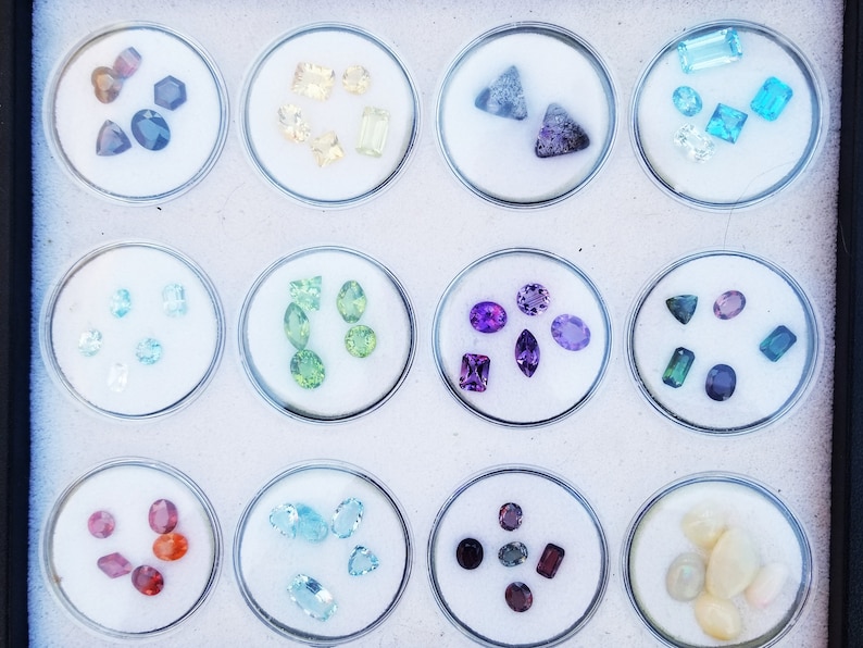 May include: A collection of 12 small glass containers, each filled with a variety of colorful gemstones. The gemstones are arranged in a grid pattern, with each container containing a different type of gemstone.