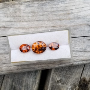 May include: Three oval-shaped gemstones in a white display box. The gemstones are orange and brown with a translucent appearance.