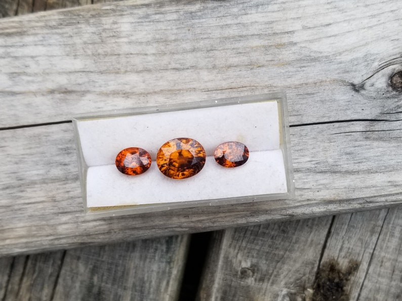 May include: Three oval-shaped gemstones in a white display box. The gemstones are orange and brown with a translucent appearance.