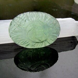 Pode incluir: Uma gema oval verde esculpida com um design floral detalhado. A gema est&aacute; apoiada em um cristal de selenita branco.