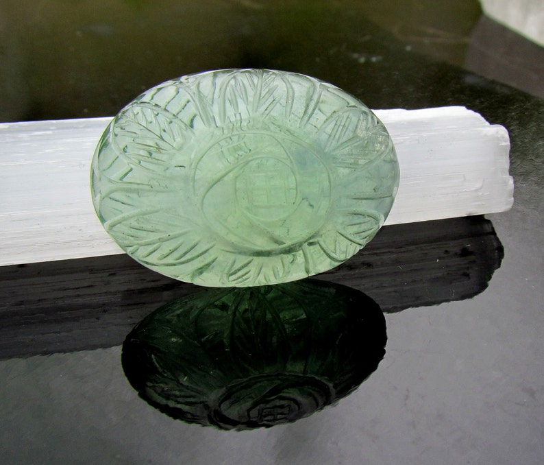 Pode incluir: Uma gema oval verde esculpida com um design floral detalhado. A gema est&aacute; apoiada em um cristal de selenita branco.