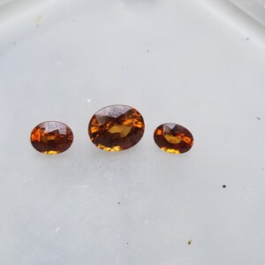 May include: Three oval-shaped gemstones, ranging in size, with a warm orange-brown hue. The gemstones are displayed on a white surface.