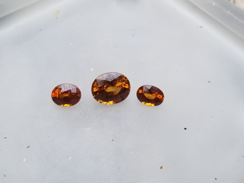 May include: Three oval-shaped gemstones, ranging in size, with a warm orange-brown hue. The gemstones are displayed on a white surface.