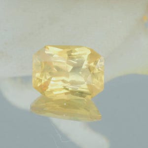 K&ouml;nnte beinhalten: Ein facettierter, rechteckig geschliffener gelber Edelstein mit reflektierender Oberfl&auml;che. Der Edelstein hat eine helle, goldgelbe Farbe und scheint auf einer reflektierenden Oberfl&auml;che zu liegen. Der Edelstein wird wahrscheinlich in Schmuck verwendet.