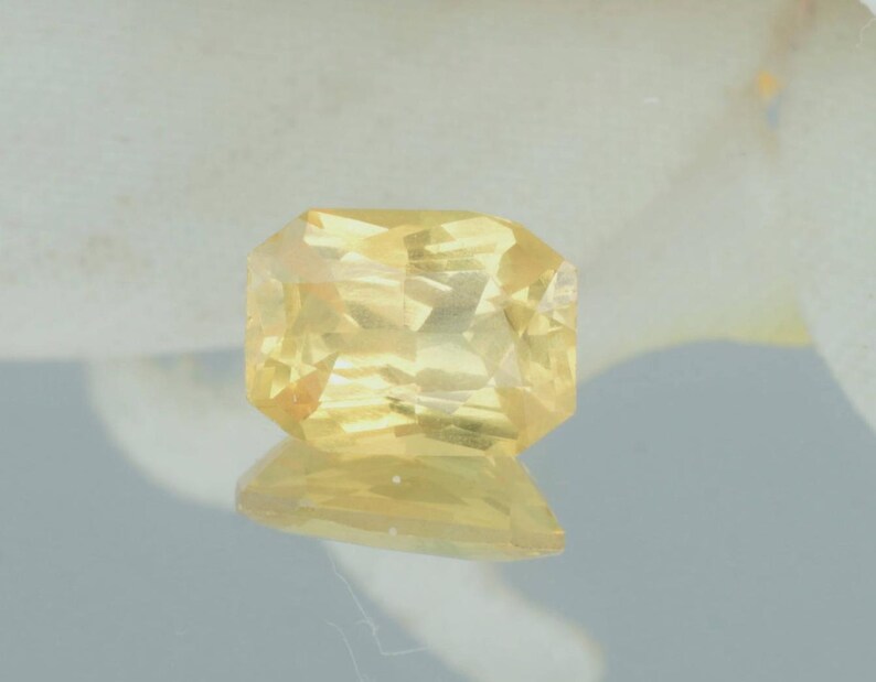 K&ouml;nnte beinhalten: Ein facettierter, rechteckig geschliffener gelber Edelstein mit reflektierender Oberfl&auml;che. Der Edelstein hat eine helle, goldgelbe Farbe und scheint auf einer reflektierenden Oberfl&auml;che zu liegen. Der Edelstein wird wahrscheinlich in Schmuck verwendet.