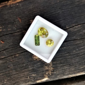 May include: Three green gemstones in a white square box. The gemstones are different shapes and sizes. The box is on a dark brown wooden surface.