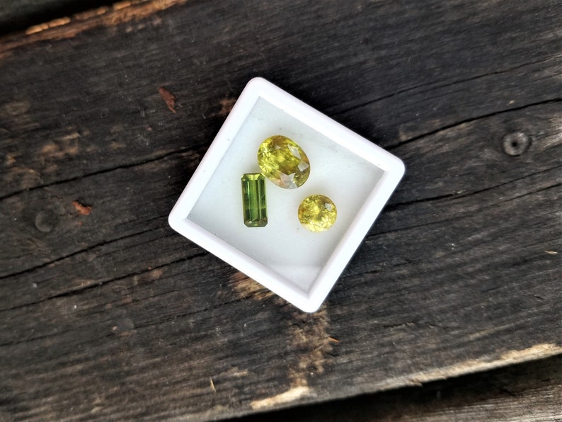 May include: Three green gemstones in a white square box. The gemstones are different shapes and sizes. The box is on a dark brown wooden surface.