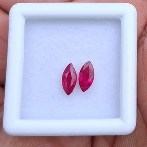 May include: Two loose marquise-cut rubies with a deep red colour and faceted surfaces, displayed side-by-side within a square white plastic display case featuring a white foam insert for secure gemstone storage and presentation.