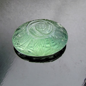 Pode incluir: Um caboch&atilde;o de pedra preciosa verde esculpido com um desenho floral. O caboch&atilde;o &eacute; redondo e tem uma superf&iacute;cie lisa e polida.