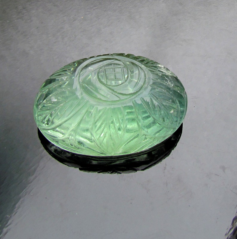 Pode incluir: Um caboch&atilde;o de pedra preciosa verde esculpido com um desenho floral. O caboch&atilde;o &eacute; redondo e tem uma superf&iacute;cie lisa e polida.