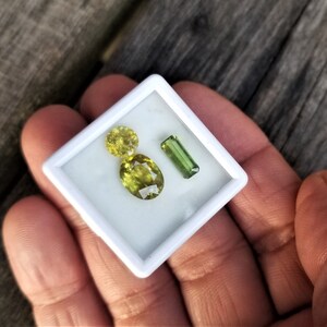 May include: Three gemstones in a white box. Two are yellow-green oval-shaped stones and one is a green rectangular stone.