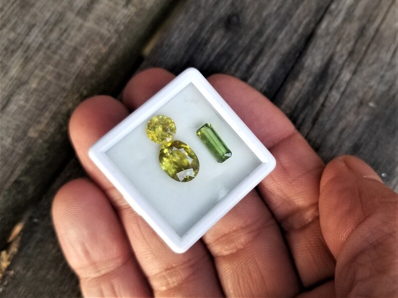 May include: Three gemstones in a white box. Two are yellow-green oval-shaped stones and one is a green rectangular stone.