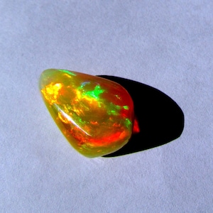 May include: A smooth, teardrop-shaped opal gemstone with a vibrant display of red, yellow, and green iridescent colors.