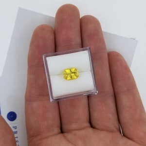 K&ouml;nnte beinhalten: Ein rechteckiger, gelber Edelstein in einer transparenten Kunststoffbox, gehalten in einer offenen Hand. Der Edelstein hat eine leuchtend gelbe Farbe. Der Hintergrund ist wei&szlig;, mit einem teilweise sichtbaren blau-wei&szlig;en Dokument.