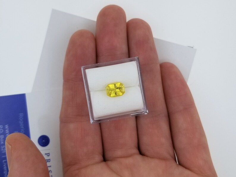 K&ouml;nnte beinhalten: Ein rechteckiger, gelber Edelstein in einer transparenten Kunststoffbox, gehalten in einer offenen Hand. Der Edelstein hat eine leuchtend gelbe Farbe. Der Hintergrund ist wei&szlig;, mit einem teilweise sichtbaren blau-wei&szlig;en Dokument.