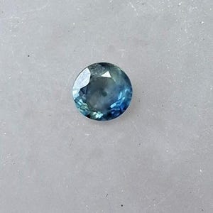 Peut inclure: Une pierre précieuse ronde à facettes présentant un dégradé de teintes bleu profond et vert sarcelle. La pierre présente une taille brillant avec de multiples facettes triangulaires qui reflètent la lumière, créant un aspect translucide.