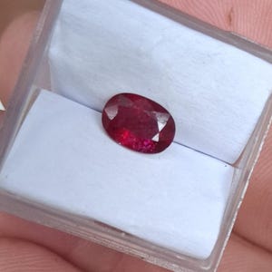 Puede incluir: Una piedra preciosa de color rojo intenso con forma ovalada y facetas, presentada dentro de una pequeña caja de almacenamiento cuadrada de plástico transparente con un inserto de espuma blanca. La piedra muestra un tono carmesí vibrante.