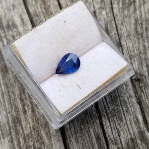Puede incluir: Una piedra preciosa de zafiro en forma de pera en una peque&ntilde;a caja blanca. El zafiro es de color azul oscuro.
