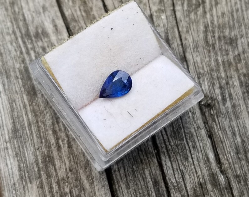 Puede incluir: Una piedra preciosa de zafiro en forma de pera en una peque&ntilde;a caja blanca. El zafiro es de color azul oscuro.