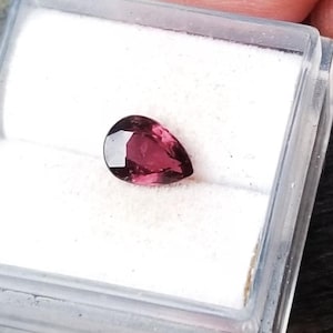 May include: A pear-shaped, deep pink gemstone in a white, square, plastic box.