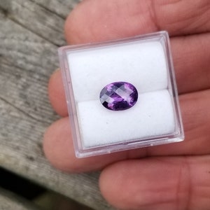 May include: A purple oval gemstone in a clear plastic box