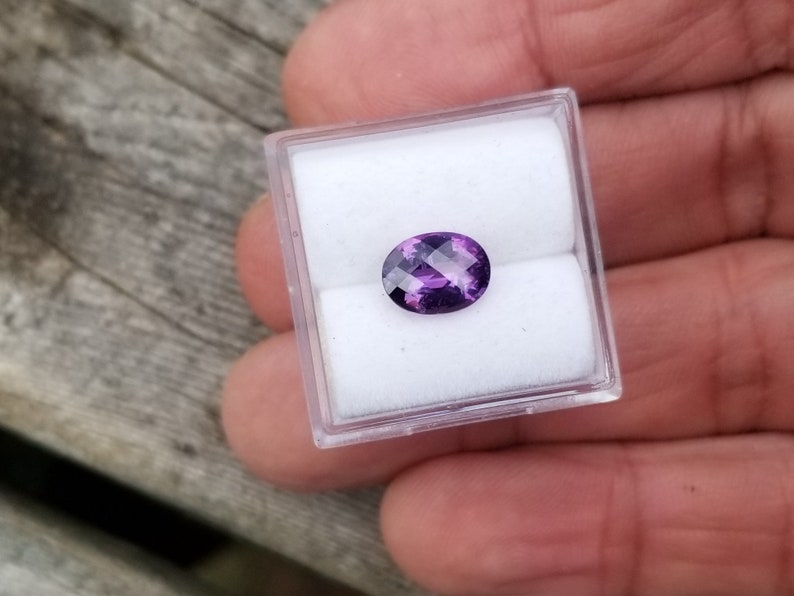 May include: A purple oval gemstone in a clear plastic box